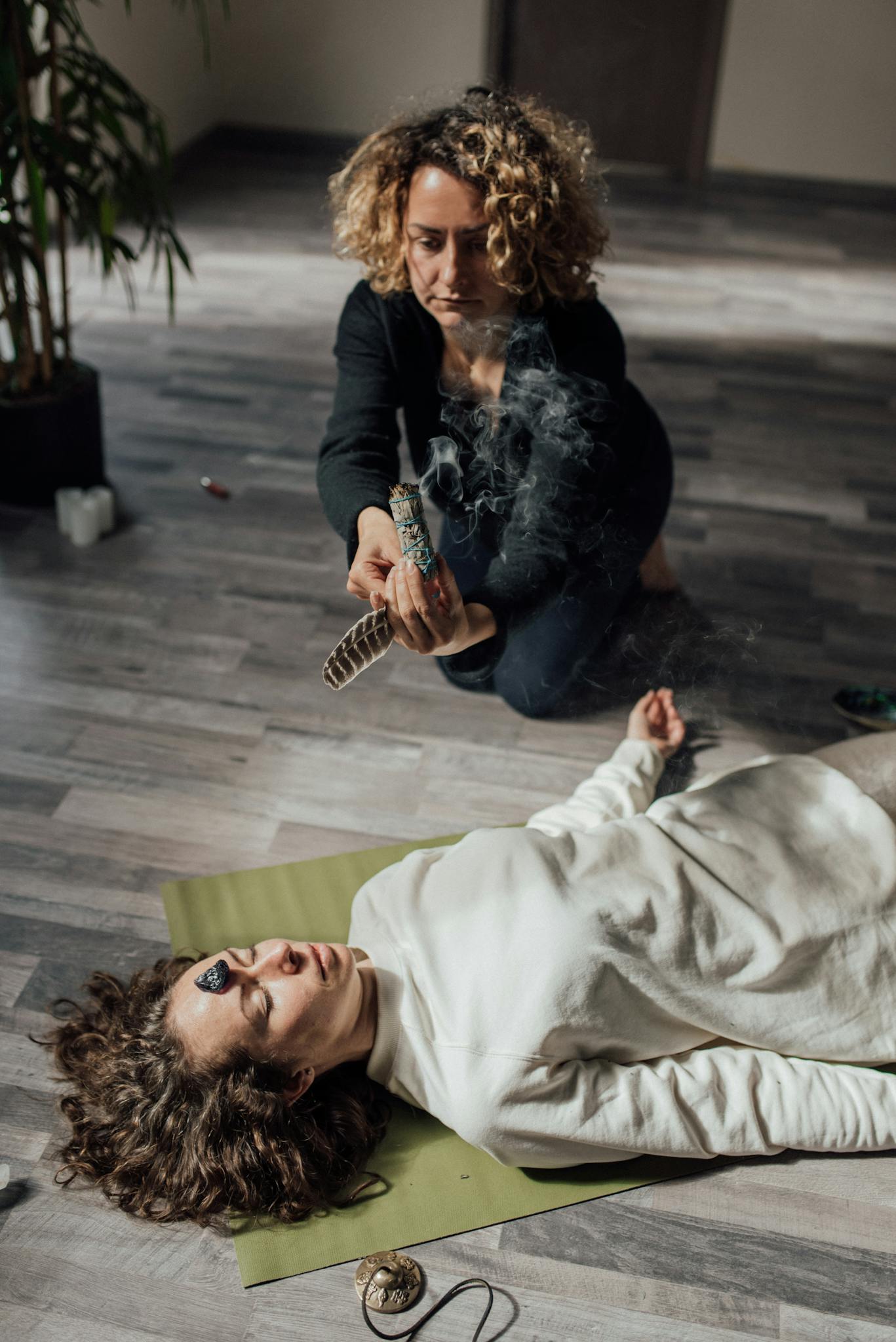 A woman performs a healing ritual with smudging and crystals on another woman indoors.